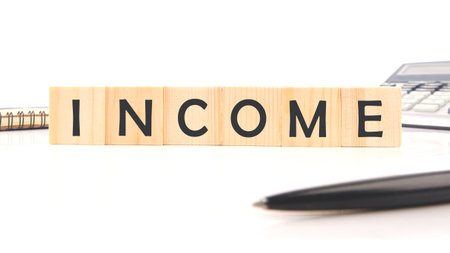 Wooden blocks spell out INCOME. A calculator, notepad, and pen sit on a white desk, suggesting planning and financial consideration for the future.の写真素材