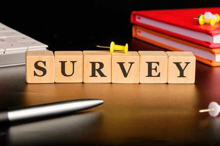 Wooden blocks spell out SURVEY. The blocks are arranged on a desk near books, a pen, a keyboard, and colorful pushpins. The surface reflects the light.の写真素材