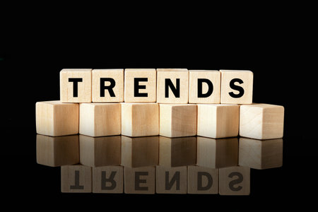 Seven wooden letter blocks stacked on a shiny, reflective surface spell out the word TRENDS against a plain black background. The reflection mirrors the word below.の写真素材