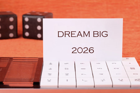 A white card displaying the words DREAM BIG 2026 is propped up on a calculator, with two black dice...