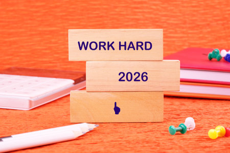 Wooden blocks stacked on an orange surface spell out Work Hard and 2026, surrounded by a pencil,...