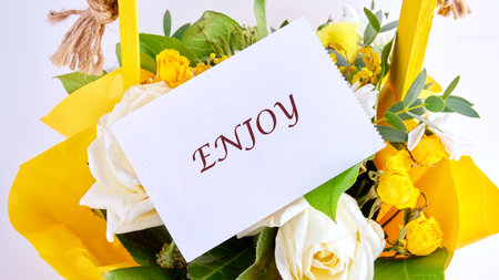 A flower arrangement is presented with white and yellow roses, green leaves, and a note card with...