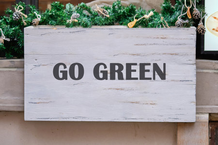 A weathered white wooden sign displays the phrase GO GREEN in bold, dark letters, topped with festive garland and pine cones. It appears during the daytime.の写真素材