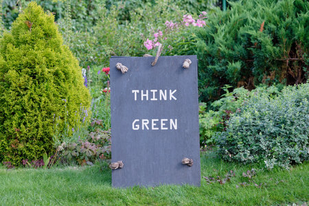 A dark Slate Board with THINK GREEN in white letters stands on green grass. Lush plants and flowers fill the background, promoting an eco-friendly message.の写真素材