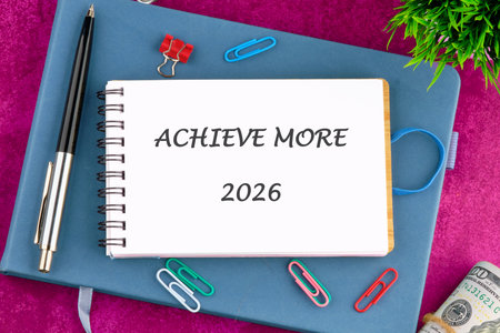 A planner with ACHIEVE MORE and 2026 displayed sits atop a blue journal. A pen, paperclips, cash, and a small plant surround it, suggesting a goal setting.の写真素材
