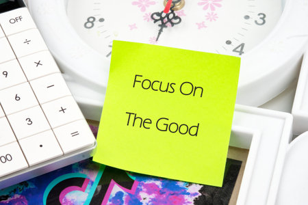 A neon yellow sticky note boldly states Focus On The Good, positioned among a white calculator, a clock face, and picture frames, suggesting a moment of positive reflection.の写真素材