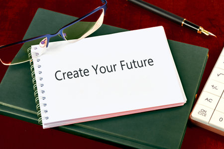 Scene features a notebook with Create Your Future printed on it. A pen, glasses and calculator also rest on the surface of a wooden desk, next to a green book.の写真素材