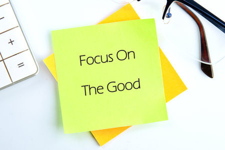 A lime green sticky note with the words Focus On The Good rests atop a yellow one, next to a calculator and glasses on a white surface, promoting a positive outlook.の写真素材