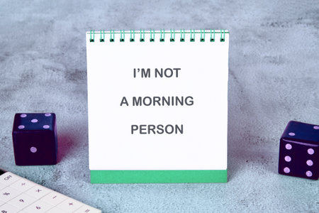 A notepad displays IM NOT A MORNING PERSON. Two dice and a calculator surround the notepad on a gray...
