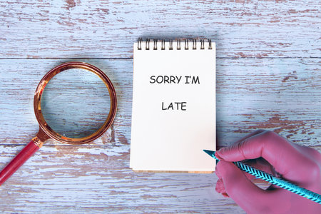 Note pad displays SORRY I AM LATE on a light blue wood table with magnifying glass. Hand holds...