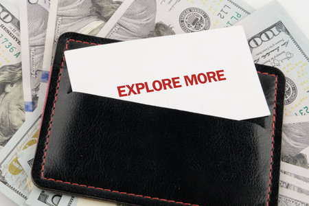 A black leather wallet rests on top of numerous United States currency bills. A card reading EXPLORE MORE is partially visible, sticking out from the wallets pocket.の写真素材