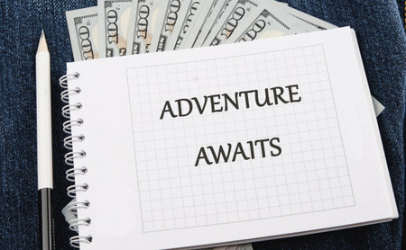 A notebook with the words ADVENTURE AWAITS rests on denim, accompanied by a pencil and some cash,...