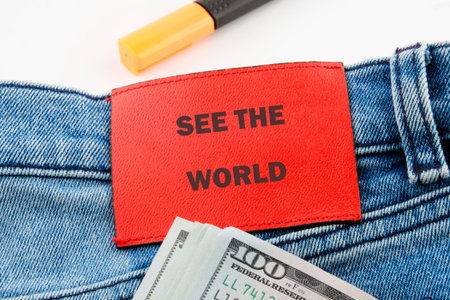 A pair of denim jeans sits with a red label that says SEE THE WORLD. A stack of United States currency and a yellow highlighter are placed beside them, suggesting travel planning.の写真素材