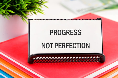 A white card displays the words PROGRESS NOT PERFECTION. The card rests on several colorful books and sits next to a small green plant. Motivation is visible.の写真素材