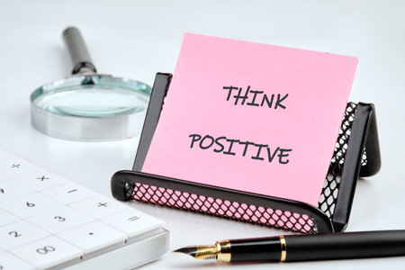 A pink sticky note propped in a black holder says THINK POSITIVE. Nearby, a calculator, pen, and magnifier suggest financial planning and a focus on achieving goals in an office setting.の写真素材