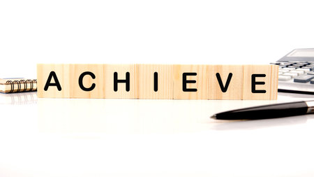 Wooden blocks spell out the word ACHIEVE near a calculator, notebook and pen, representing success and goals in business or academics, on a bright white surface.の写真素材