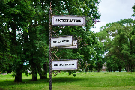 In a leafy park, an ornate metal sign with three duplicate plaques spells out PROTECT NATURE. It...