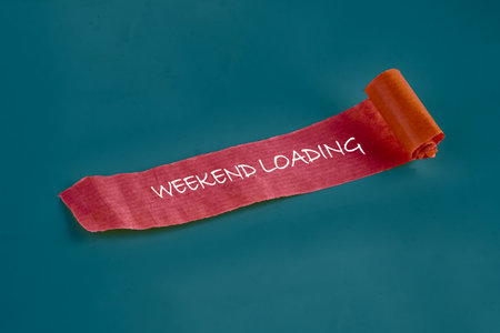 A rolled piece of orange paper displaying the phrase WEEKEND LOADING is staged over a calming teal background, alluding to awaiting the joys of days away from work.の写真素材