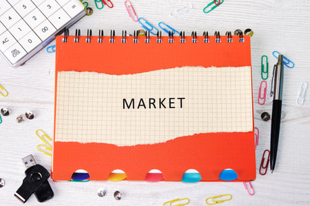 Orange notebook with the word Market written on it rests on a wooden desk among calculator, pen, paper clips, usb drive, and thumb tacks. Planning concept is the goal.の写真素材