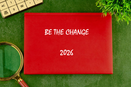 A red book sits with the text BE THE CHANGE 2026, a calculator, a magnifying glass, and a small...