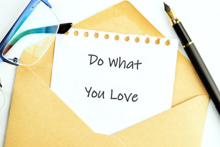 A note with Do What You Love sits inside an envelope. A fountain pen and eyeglasses are placed beside it, ready for writing, on a white table.の写真素材