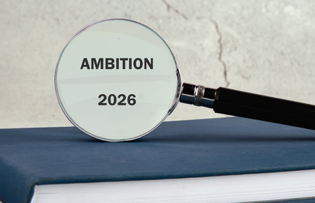 A magnifying glass highlighting the word ambition alongside the year 2026, resting on a blue book....