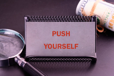 A sleek business card holder displays the phrase PUSH YOURSELF prominently. Nearby, rolled currency hints at financial success. This setup inspires ambition and self-improvement.の写真素材