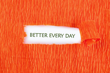 Against a vivid orange background, a piece of paper peeks through, revealing the uplifting message of better every day. This design encourages continuous self-improvement.の写真素材