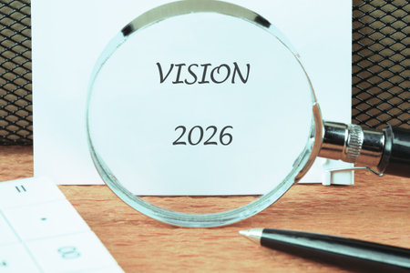 A magnifying glass enlarges the words VISION 2026. A pen and calculator sit on a wood-grain surface,...