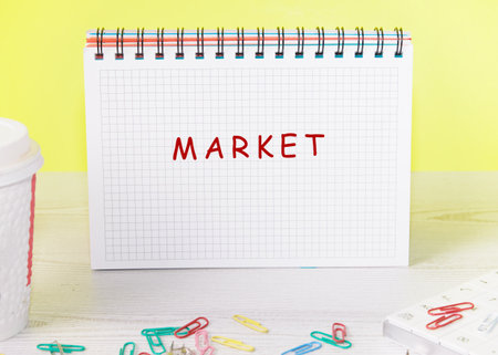 An open, spiral-bound notebook sits on a desk with colorful paperclips scattered around. The word Market is handwritten on the notepad against a bright yellow background.の写真素材
