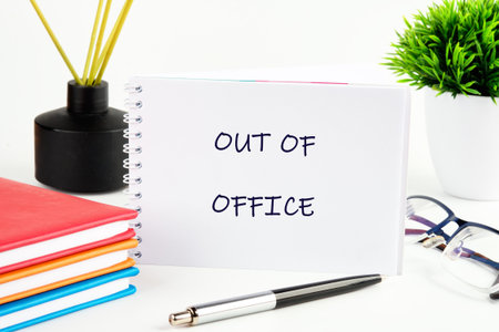 A note card displays the words OUT OF OFFICE sitting a pen, books, glasses, a plant, and an aromatherapy diffuser on a bright white desk.の写真素材