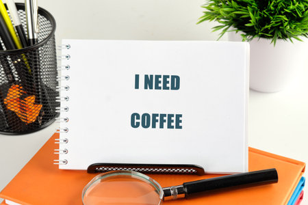 A spiral notebook on top of orange books displays the phrase I NEED COFFEE. A magnifier, pen holder, and plant are nearby on a white surface.の写真素材