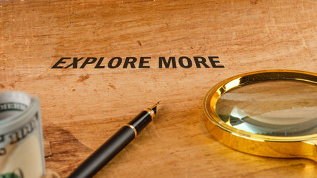 A golden magnifying glass, rolled United States currency, and a fountain pen sit on a desk next to the inscription explore more. This evokes adventure and learning.の写真素材