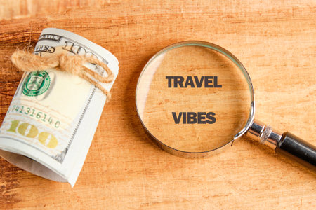 A stack of one hundred dollar bills secured with twine sits beside a magnifying glass that shows the words TRAVEL VIBES on a textured wooden surface.の写真素材