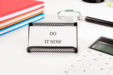 A card reading DO IT NOW sits on a desk with stacked colorful books, a magnifying glass, and a calculator, promoting instant task completion.の写真素材