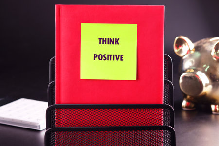A bright red notebook with a THINK POSITIVE sticky note sits upright in a black wire organizer. A shiny golden piggy bank stands nearby on a dark surface.の写真素材