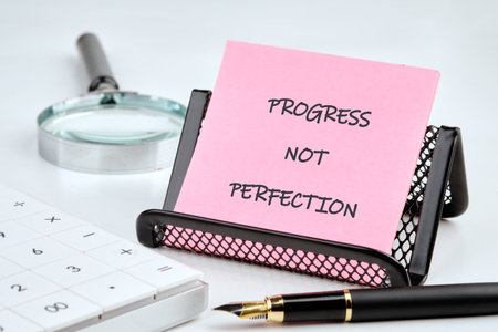Pink notepad displays the words PROGRESS NOT PERFECTION. Calculator, pen, and magnifying glass are nearby. The scene speaks of improvement, focus, and striving forward.の写真素材