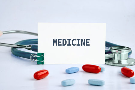 A white card with the word MEDICINE stands near a stethoscope and an assortment of red and blue pills on a white surface. It suggests healthcare and treatment.の写真素材