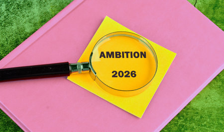 A magnifying glass focuses on the words Ambition 2026 written on a yellow sticky note. The note sits...