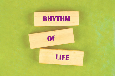Three light-colored wooden blocks are stacked. Each block has a word in purple letters forming the phrase RHYTHM OF LIFE on a bright, textured, green background.の写真素材