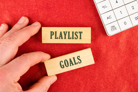 A hand arranges wooden blocks reading PLAYLIST and GOALS on a red surface, next to a calculator....