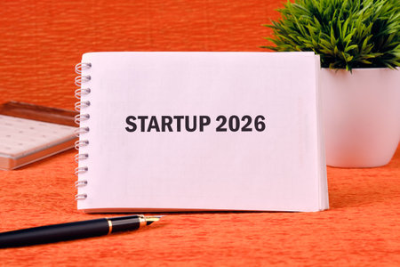 A notebook displays the words STARTUP 2026 next to a pen, desk calendar, and potted plant. The orange surface suggests preparation for future ventures and growing prosperity.の写真素材
