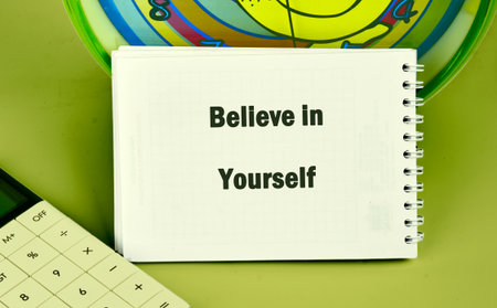 A white notepad displays the words Believe in Yourself, resting next to a calculator and a vibrant clock.の写真素材