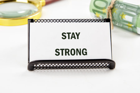 A small sign displays the uplifting message stay strong set against a backdrop of money and a magnifying glass, symbolizing determination and focus in challenging times.の写真素材