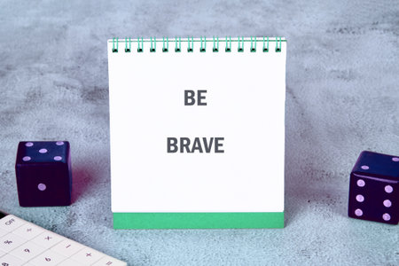 A motivational note stands upright on a desk, boldly stating the words BE BRAVE. Two colorful dice sit nearby, enhancing a creative workspace atmosphere filled with inspiration.の写真素材