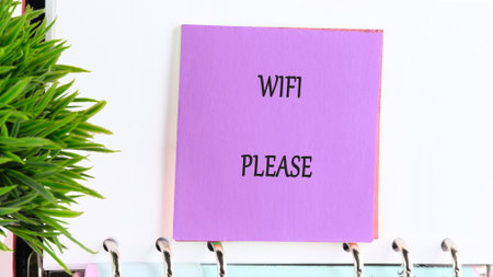 A square lavender note requests internet connectivity with the words WIFI PLEASE. The note is clipped to a notebook and sits beside a lush, green plant.の写真素材