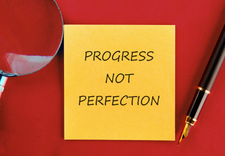 A yellow sticky note displaying the text PROGRESS NOT PERFECTION rests on a red surface, accompanied by a magnifying glass and a fountain pen.の写真素材