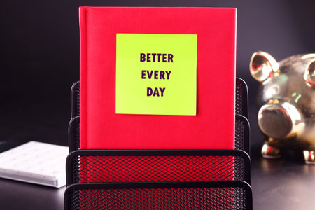 A red book rests in a metal organizer with a neon yellow sticky note that reads BETTER EVERY DAY. Nearby is a golden piggy bank on a dark surface, suggesting a focus on financial goals.の写真素材