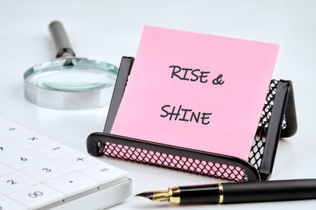 A pink note displays RISE SHINE next to a pen, calculator, and magnifying glass. It suggests a bright start, possibly for planning or problem-solving tasks at the office.の写真素材