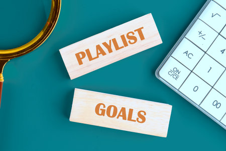 Flat lay of wooden blocks with the words Playlist and Goals, a calculator, and magnifying glass sit atop a teal background, perhaps for planning how to make the perfect songs list.の写真素材